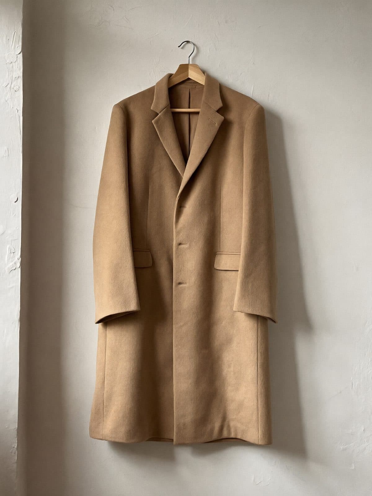 A camel wool coat on a wooden hanger against a warm off-white wall.