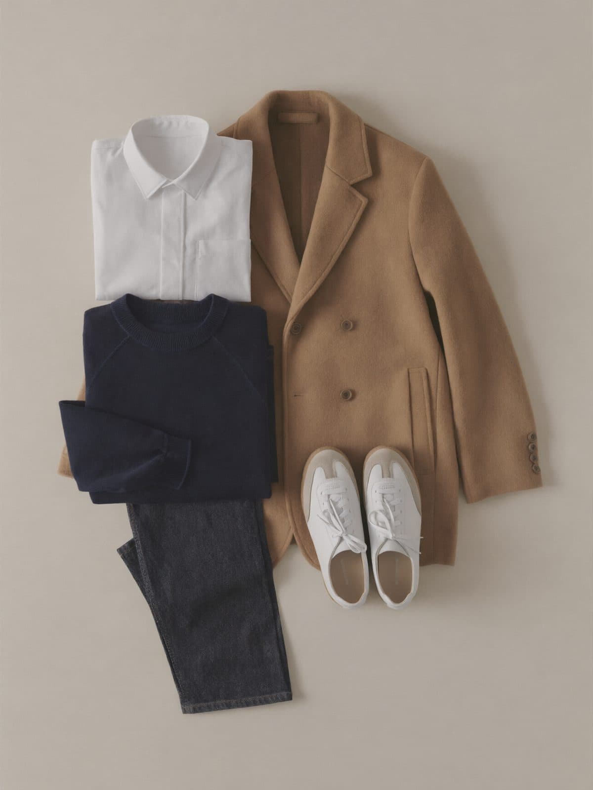 A flat lay of a small capsule wardrobe: dark jeans, a white shirt, a navy sweater, a camel coat and white sneakers.