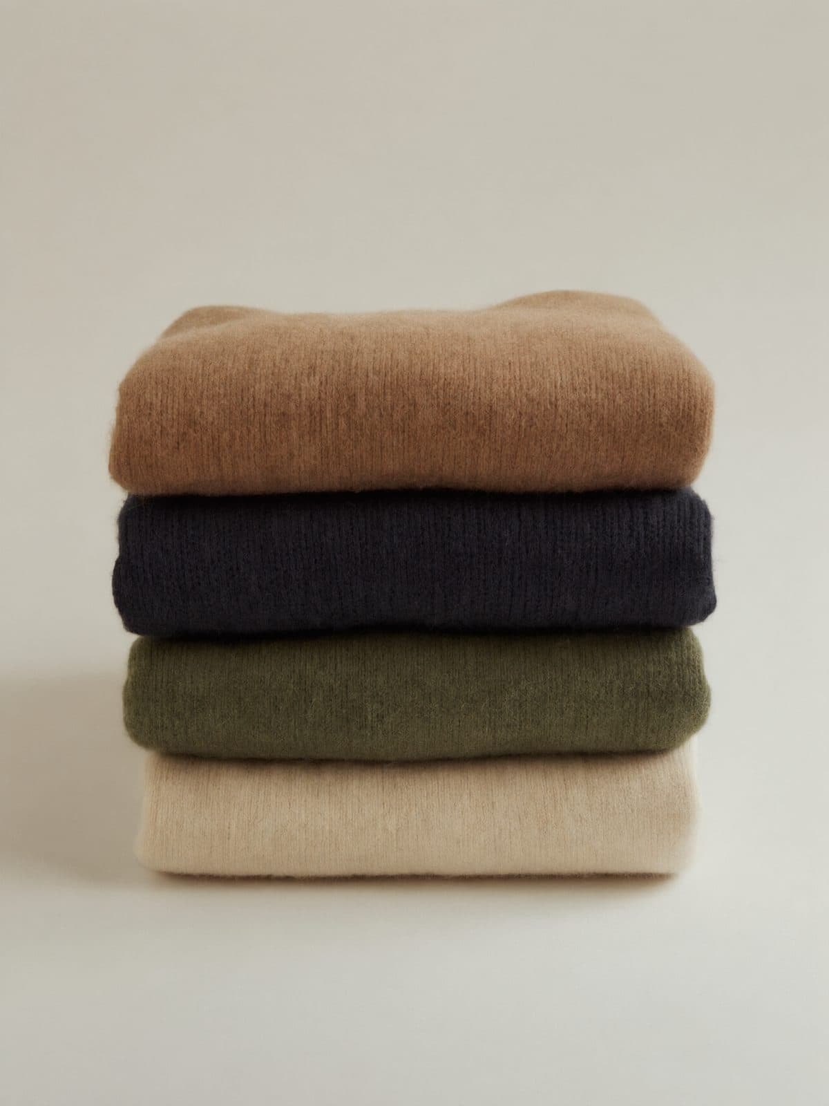Folded knitwear in camel, navy, olive and cream stacked on light grey paper.
