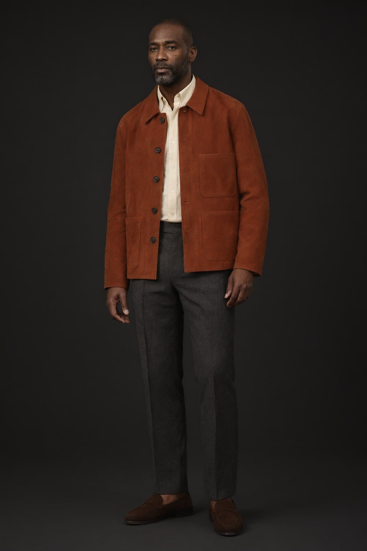 A man wears the rust suede jacket over a cream shirt with charcoal trousers and brown suede loafers.
