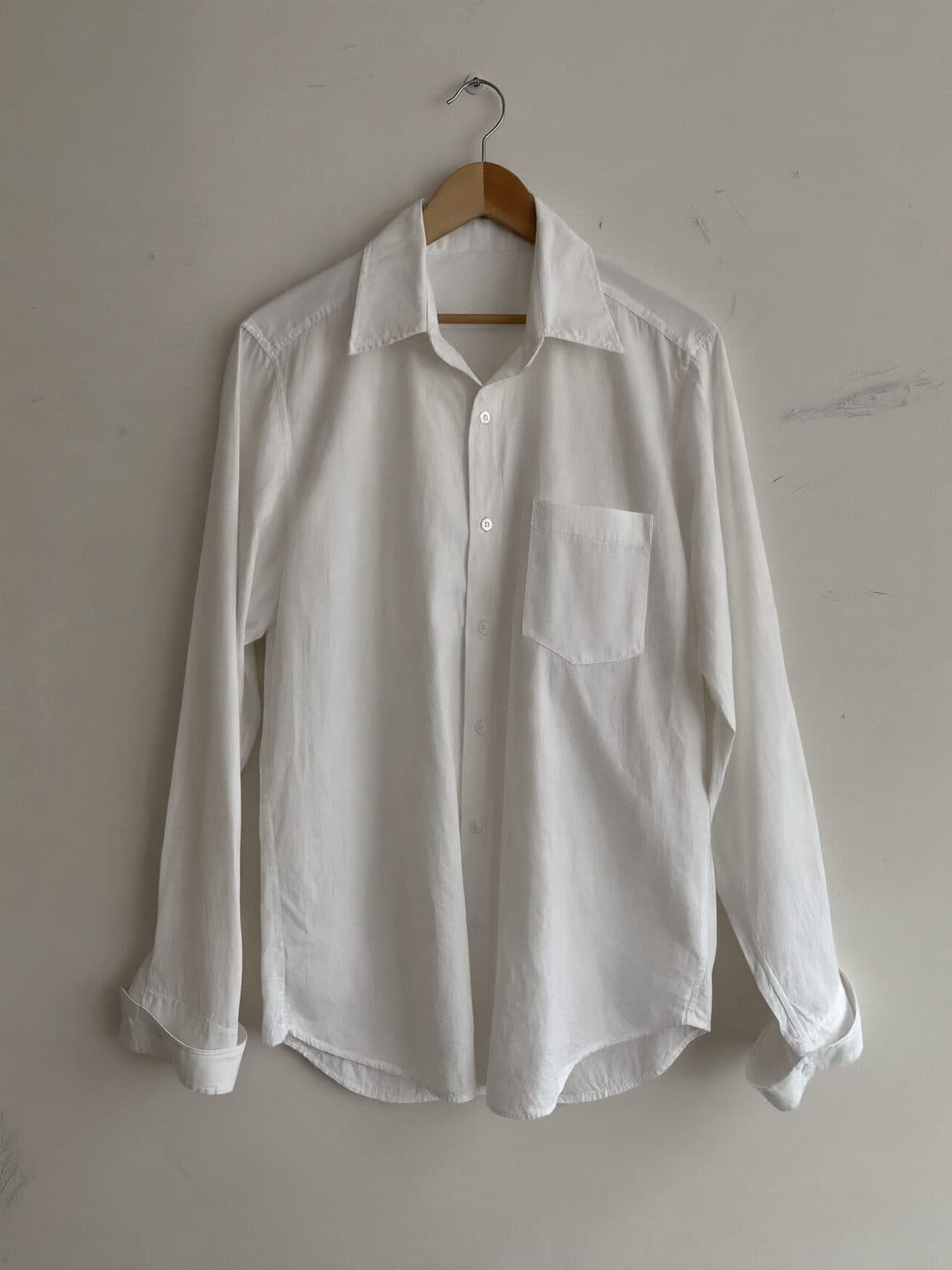 A crisp white cotton shirt on a wooden hanger against a light grey wall.